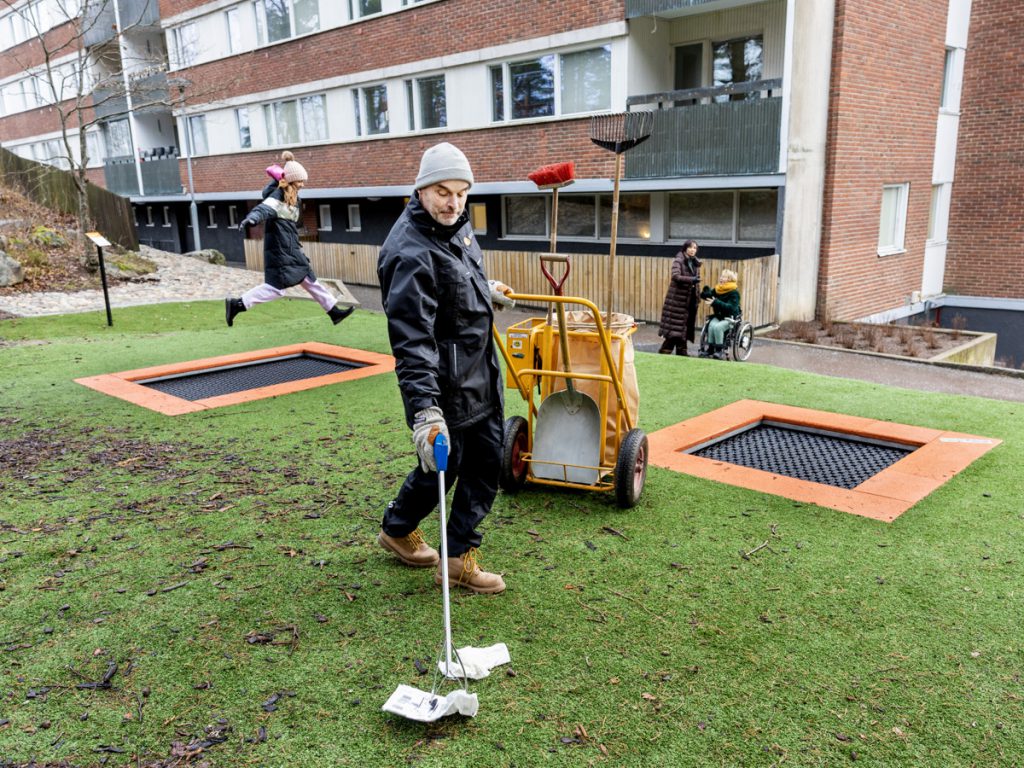 Fastighetsskötare plockar skräp, barn hoppar på studsmatta och två kivnnor pratar med varandra i bakgrunden. 